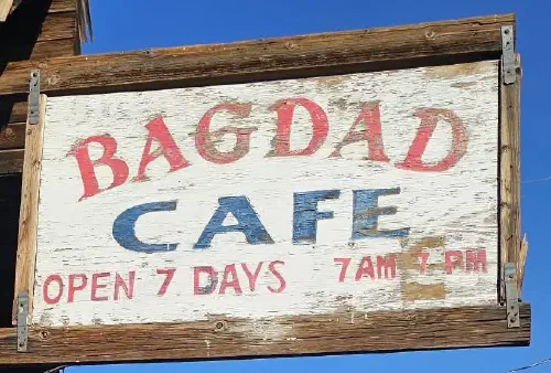 Enseigne en bois délavé du Bagdad Café, « Open 7 days, 7AM 7PM », sur fond de ciel bleu