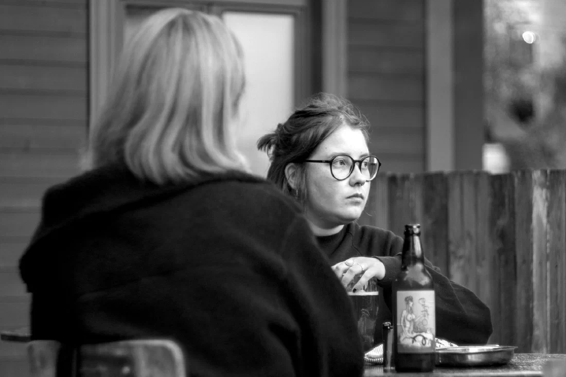Scène de terrasse en noir et blanc à Rīga : une jeune femme brune aux lunettes rondes et piercing au nez, l'air pensif, assise face à une femme blonde vue de dos ; sur la table, une bouteille de bière artisanale à l'étiquette illustrée.