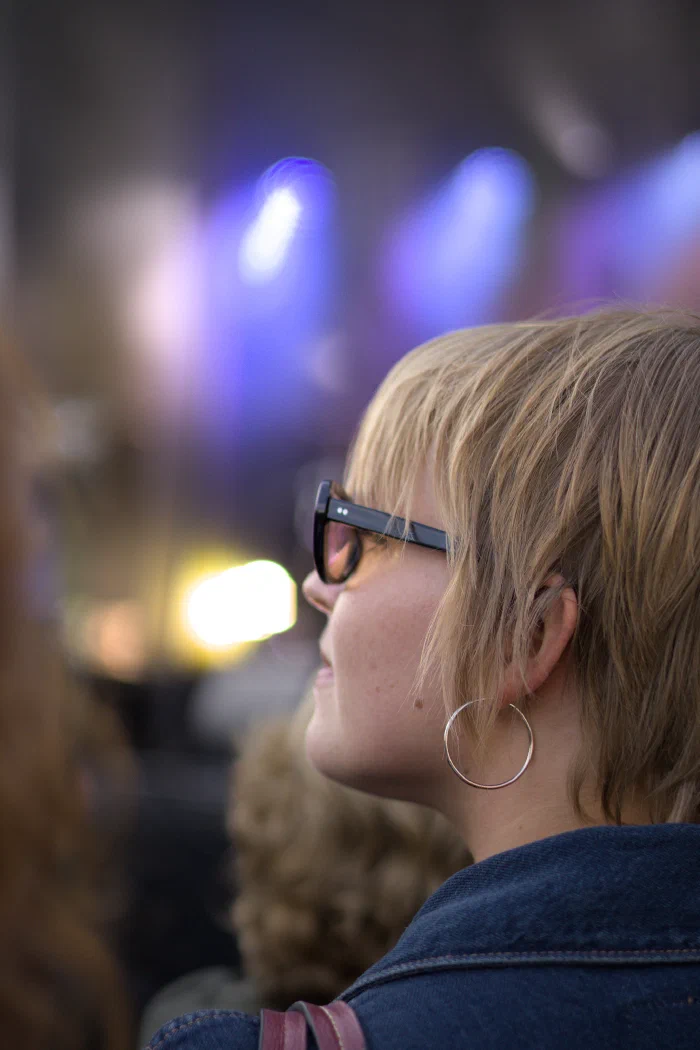 Color profile portrait of a short-haired blonde young woman, black-framed glasses and a large silver hoop earring, looking toward the stage; blurred blue and yellow lights in the background. Binic Folks Blues Festival 2024.