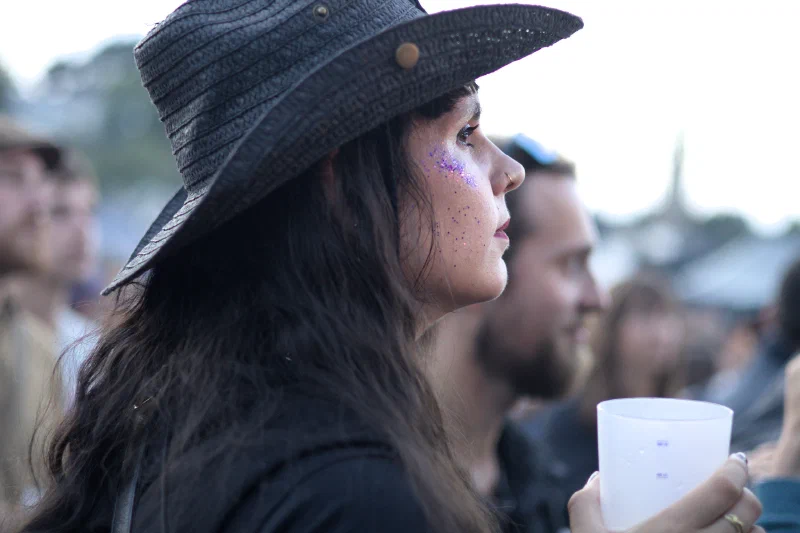 Color profile portrait of a dark-haired young woman wearing a black straw hat in a festival crowd, purple glitter on her cheek, a white cup in hand, blurred and luminous background. Binic Folks Blues Festival 2025.