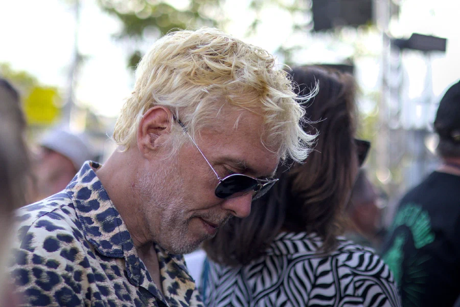 Portrait de profil d'un homme aux cheveux décolorés blond platine, barbe grisonnante, lunettes d'aviateur et chemise léopard, tête baissée dans la foule d'un festival en plein jour ; arrière-plan flou de scène et de feuillages.
