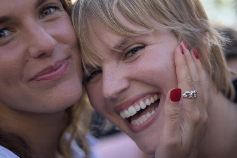 Portrait rapproché en couleur de deux jeunes femmes blondes joue contre joue en plein jour ; celle de droite rit largement, une main aux ongles vernis de rouge posée sur sa joue, celle de gauche esquisse un sourire plus retenu. Festival de Binic.