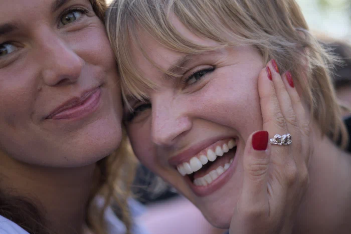 Close color portrait of two blonde young women cheek to cheek in broad daylight; the one on the right laughs widely, a hand with red-painted nails resting on her cheek, the one on the left holds a more restrained smile. Binic festival.