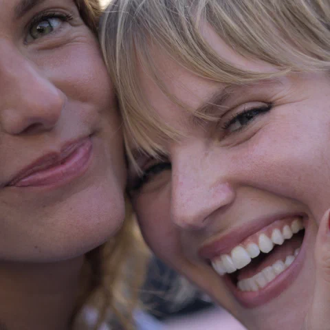 Portrait rapproché en couleur de deux jeunes femmes blondes joue contre joue en plein jour ; celle de droite rit largement, une main aux ongles vernis de rouge posée sur sa joue, celle de gauche esquisse un sourire plus retenu. Festival de Binic.