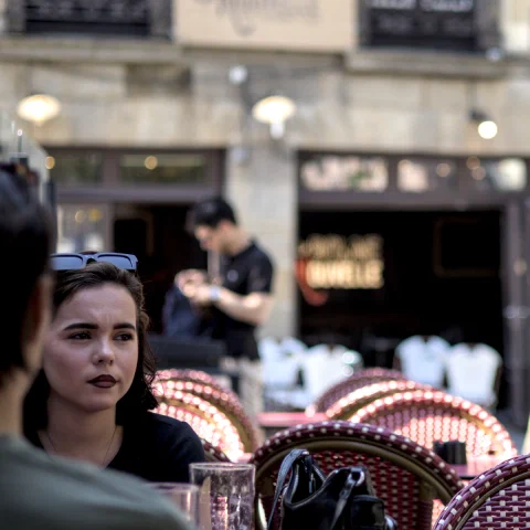 Jeune femme brune aux lèvres sombres, songeuse, à une terrasse de café à Rennes ; en arrière-plan flou, l'enseigne « La Bonne Nouvelle » et des passants.