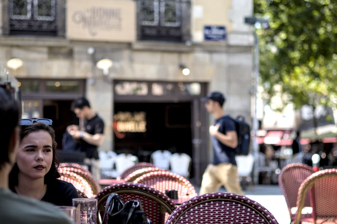 Dark-haired young woman with dark lips, pensive, at a café terrace in Rennes; blurred in the background, the sign reading 'La Bonne Nouvelle' and passers-by.