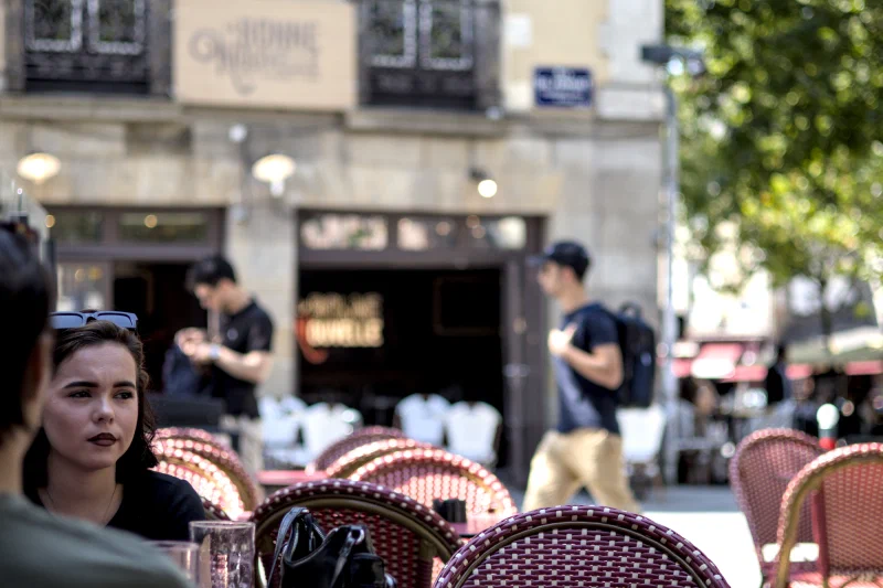 Jeune femme brune aux lèvres sombres, songeuse, à une terrasse de café à Rennes ; en arrière-plan flou, l'enseigne « La Bonne Nouvelle » et des passants.