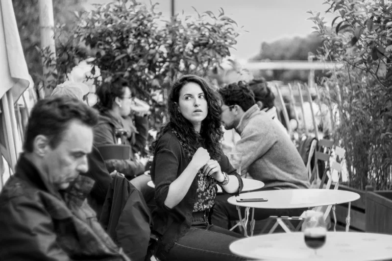 Scène de terrasse en noir et blanc à Paris : une jeune femme brune aux longs cheveux bouclés se retourne, l'air aux aguets, la main ramenée contre sa poitrine ; au premier plan flou, un homme penché en avant ; derrière, des clients attablés, des guéridons ronds, un téléphone et un verre de vin.