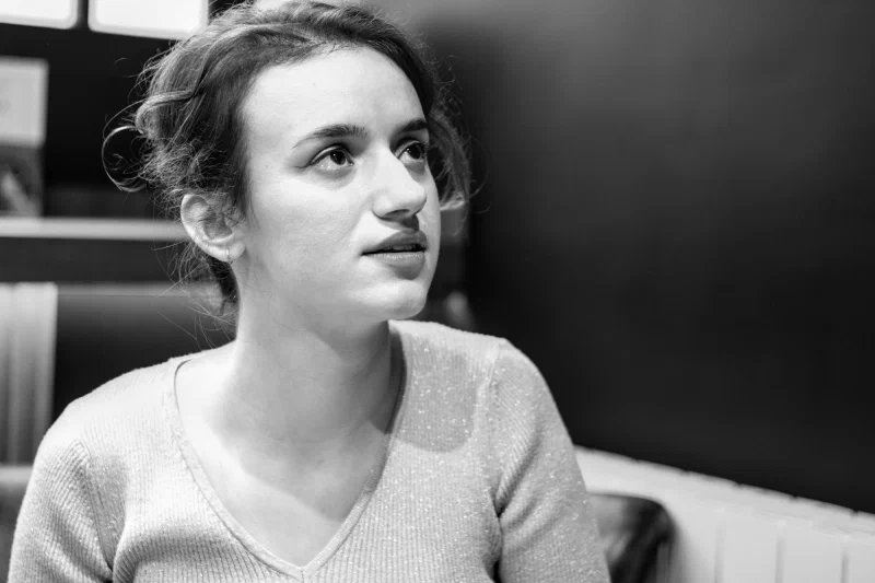 Portrait en noir et blanc de la romancière Perrine Tripier, de trois quarts, cheveux relevés, le regard porté hors-champ vers le haut, les lèvres entrouvertes, à l'écoute ; lumière douce, fond sombre.