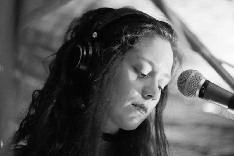 Close-up black-and-white portrait of a singer with long curly dark hair, headphones over her ears, leaning in close to a microphone, eyes closed, mid-performance. Pretplūsma concert at Aleponija, Rīga.