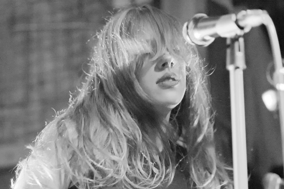 Close-up black-and-white portrait of a singer with long wavy light hair, bangs swept across her eyes, lips parted close to a mic stand. Pretplūsma concert at Aleponija, Rīga.