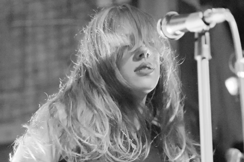 Close-up black-and-white portrait of a singer with long wavy light hair, bangs swept across her eyes, lips parted close to a mic stand. Pretplūsma concert at Aleponija, Rīga.