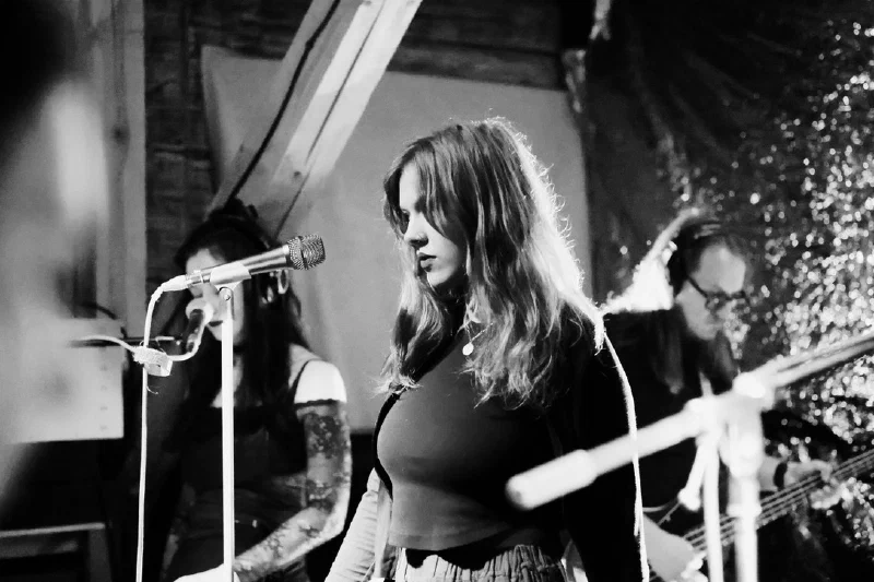 Black-and-white concert photograph: at the centre, a singer with shoulder-length hair stands near a mic stand, guitar strapped on; blurred in the background, a tattooed musician sits near a microphone on the left, a bass player with glasses on the right; a sequined backdrop behind them. Pretplūsma concert at Aleponija, Rīga.