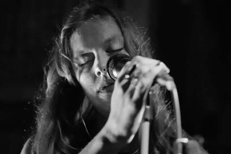 High-contrast, low-key close-up portrait of a singer with wavy hair: she grips the microphone with both hands, rings on her fingers, eyes closed, half her face in shadow. Pretplūsma concert at Aleponija, Rīga.