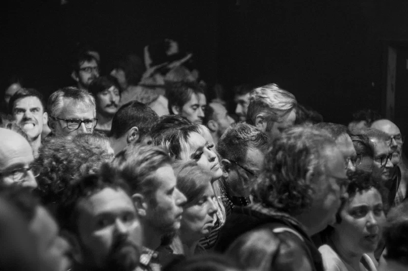 Black-and-white crowd of spectators during a concert, faces pressed close, turned toward the stage, a blonde woman at the center with half-closed eyes, deep black background.