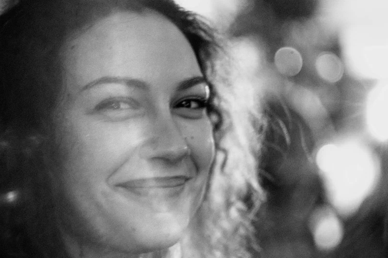 Portrait rapproché en noir et blanc d'une jeune femme souriant, le regard de côté ; la moitié droite du visage se dissout dans un halo lumineux et des cercles de bokeh, fort grain. Trans Musicales 2023, Rennes.