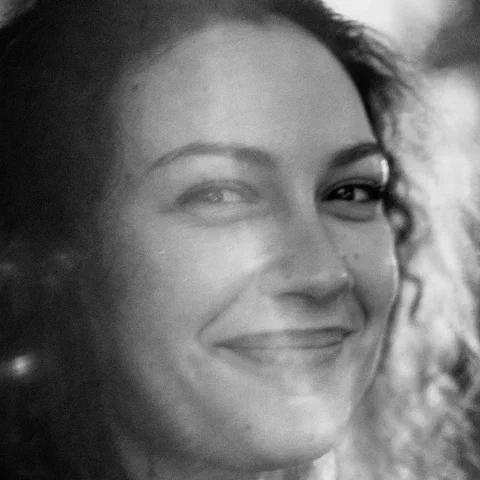 Portrait rapproché en noir et blanc d'une jeune femme souriant, le regard de côté ; la moitié droite du visage se dissout dans un halo lumineux et des cercles de bokeh, fort grain. Trans Musicales 2023, Rennes.