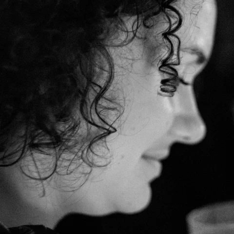 Profil en noir et blanc d'une jeune femme aux cheveux bouclés, regard baissé, dans une lumière sombre et un grain marqué ; au premier plan flou, une main tenant un gobelet. Trans Musicales 2023, Rennes.