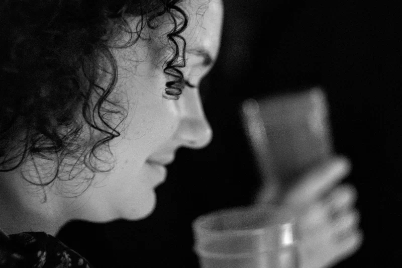 Profil en noir et blanc d'une jeune femme aux cheveux bouclés, regard baissé, dans une lumière sombre et un grain marqué ; au premier plan flou, une main tenant un gobelet. Trans Musicales 2023, Rennes.