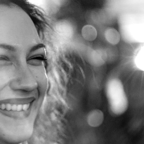 Portrait rapproché en noir et blanc d'une jeune femme aux cheveux bouclés riant largement, dents visibles, le côté droit du visage fondu dans un halo lumineux et des cercles de bokeh, fort grain. Trans Musicales 2023, Rennes.