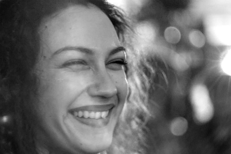 Portrait rapproché en noir et blanc d'une jeune femme aux cheveux bouclés riant largement, dents visibles, le côté droit du visage fondu dans un halo lumineux et des cercles de bokeh, fort grain. Trans Musicales 2023, Rennes.