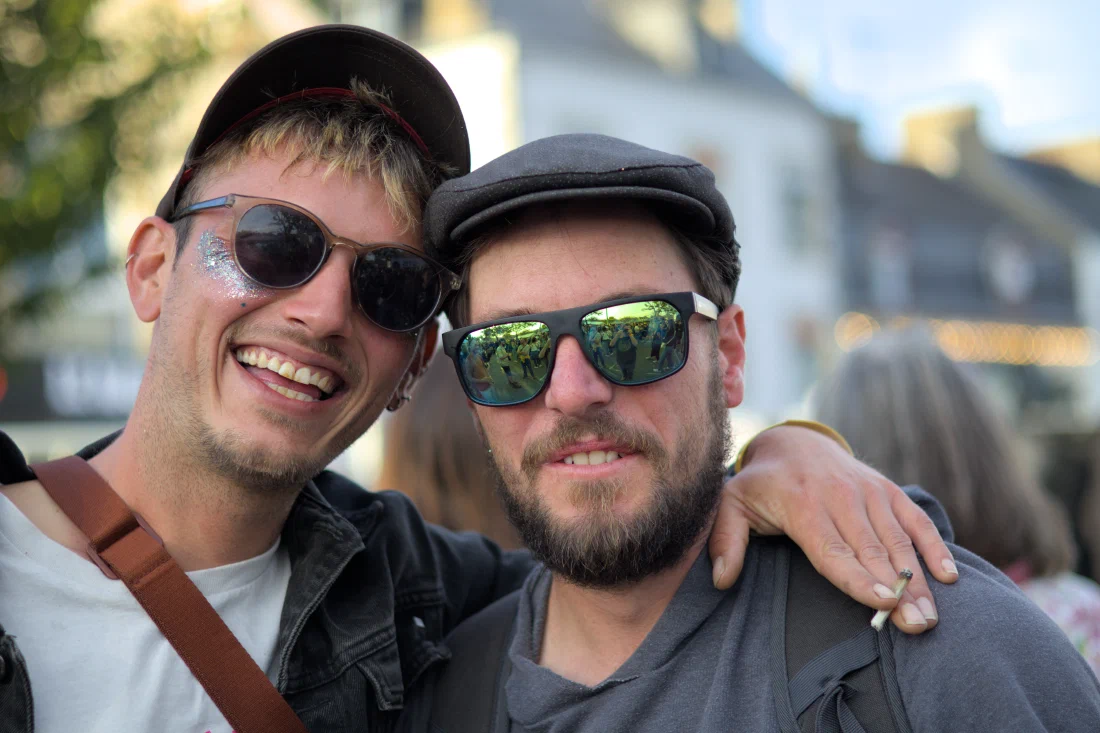 Two grinning men arm in arm, one with glitter under his eye, the other in a flat cap and mirrored sunglasses, Binic Folk Blues Festival 2024
