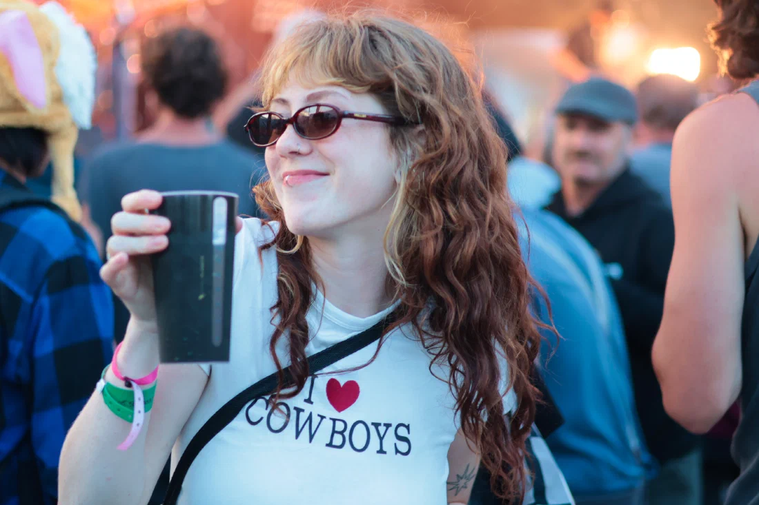 Woman in an I ♥ Cowboys tee and sunglasses raising a dark festival cup at golden hour, Binic Folk Blues Festival 2026