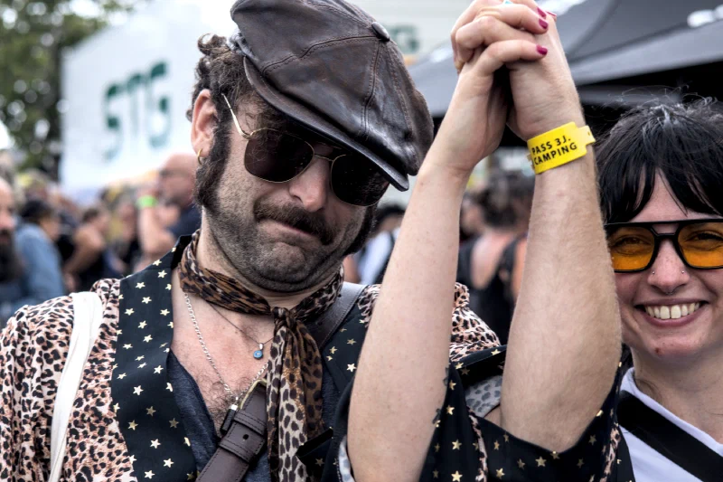 A couple with clasped hands raised in the air, the man in a flat cap and star-print vest, the woman in orange-tinted glasses, Binic Folk Blues Festival 2025