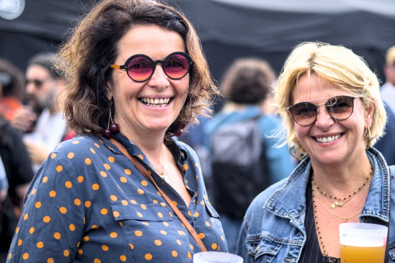 Two women laughing with beers in hand, one in round red-tinted glasses with cherry earrings, the other in a denim jacket, Binic Folk Blues Festival 2025
