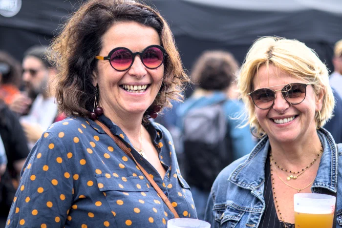 Two women laughing with beers in hand, one in round red-tinted glasses with cherry earrings, the other in a denim jacket, Binic Folk Blues Festival 2025