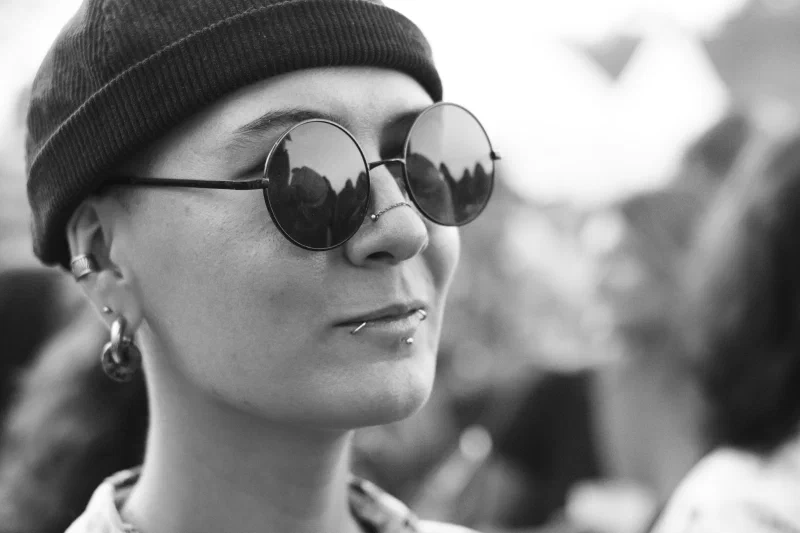 Black-and-white close-up of a festival-goer in a beanie and round mirrored sunglasses reflecting the crowd, Binic Folk Blues Festival 2026